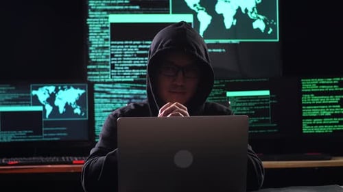 Asian Male Hacker In A Hood Looking At Camera While Works On A Laptop With Maps And Data