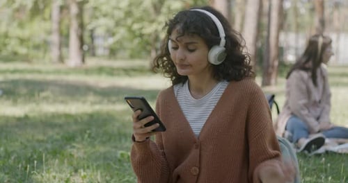 Cheerful Young Woman Using Smartphone and Listening to Music with Headphones in Park on Summer Day