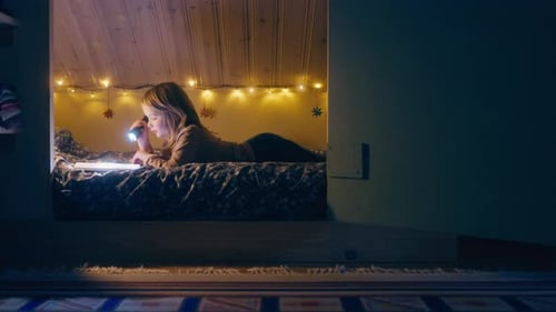 Boy lies on reading a book with flashlight in hand in whimsical nook