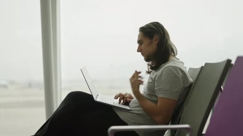Young Long Haired Man Using His Laptop Sit on an Airport Middle Shot