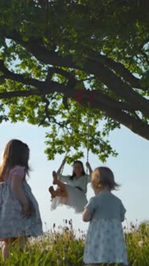Vertical of Sisters Walking Near Clean Green Field to Their Mother Swinging on Swing Tied to an Old