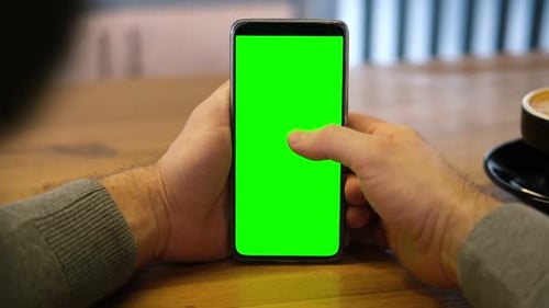 Young Man Sitting at Cafe Holding Smartphone Green Mockup Screen in Hand Male Person Using Chroma