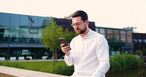 Serious businessman using on mobile phone standing on street near office building. Handsome male tex