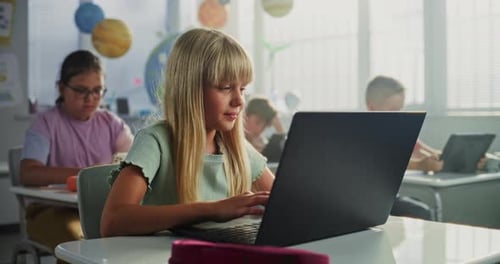 Grundschülerin sitzt am Schreibtisch und benutzt Laptop im Klassenzimmer