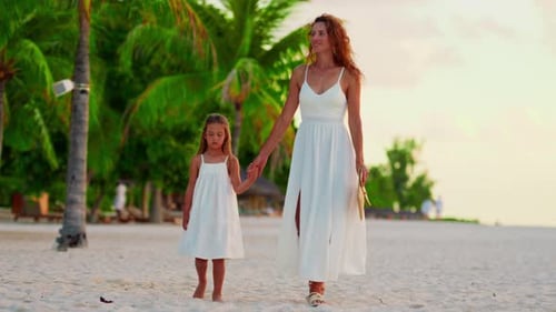A Beautiful Mother and Her Little Daughter in White Dresses Walk Hand in Hand Along the Ocean Shore