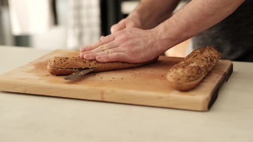 Man slicing healthy baguette on wooden chopping block on the kitchen countertop.