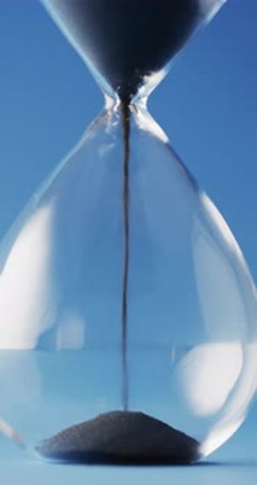 Vertical video of close up of hourglass with black sand pouring with copy space on blue background