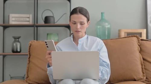 Woman uses laptop and phone sitting on couch