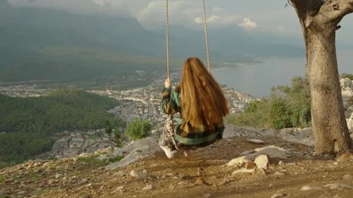 At the mountain summit, near the edge of the cliff, a young woman fearlessly swings on a swing.