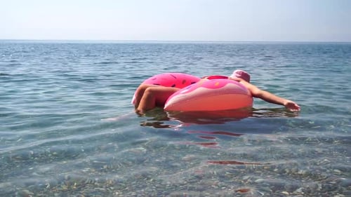 Mulher de chapéu nas férias de verão flutua em um colchão de donut inflável rosa, um anel de natação de brinquedo aquático