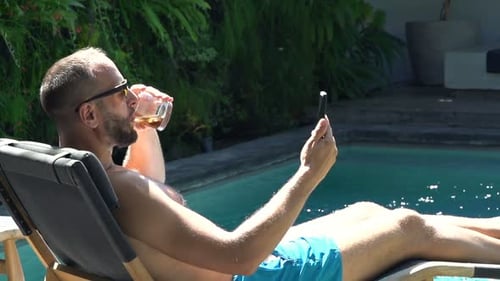 Man relaxes by pool chatting on smartphone and drinking at luxury resort