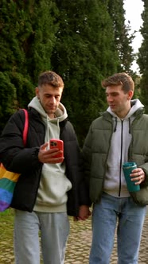 Gay couple walking in park, holding hands and using smartphone