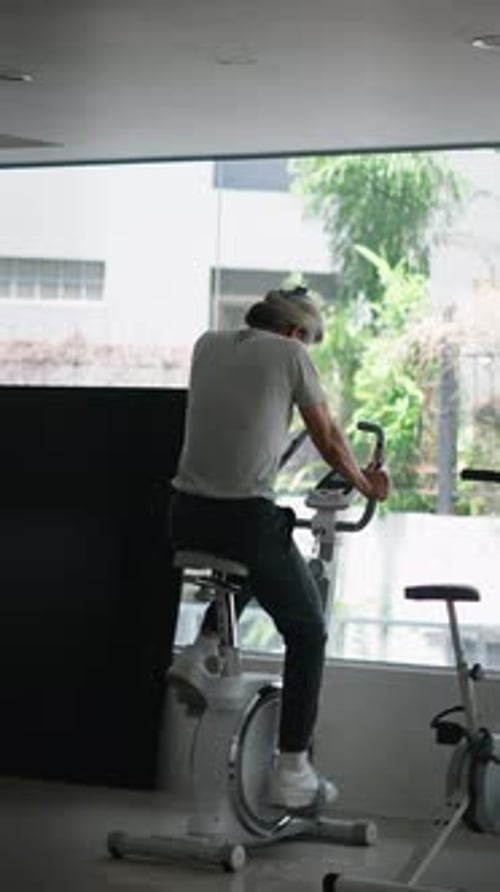 Fitness Enthusiast Pedaling on an Exercise Bike in a Large Urban Gym Overlooking a Garden