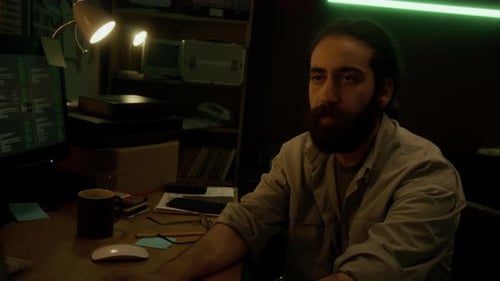 Man Sitting at Cluttered Desk, Intently Working on Computer in Dimly Lit Room