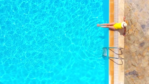 Top View of a Woman in Yellow Swimsuit and a Hat Sits on the Edge of the Pool
