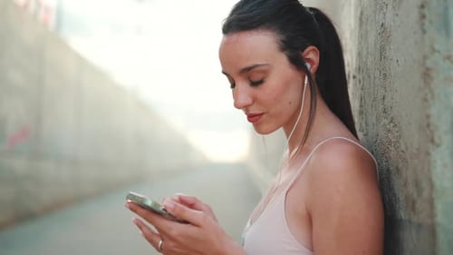 Woman Using Phone With Earbuds Against Urban Wall