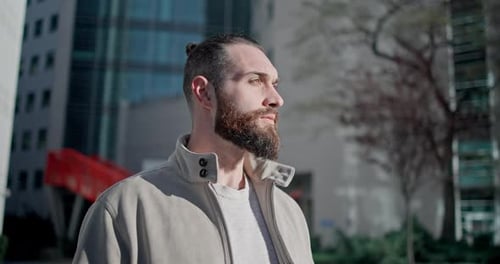 A Young Man with Stylish Trendy Hairstyle and Beard in Casual Clothes Stands on the Street in the