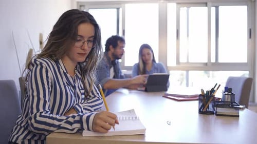 Side view of young woman taking notes and looking to camera while blurred group of two young busines