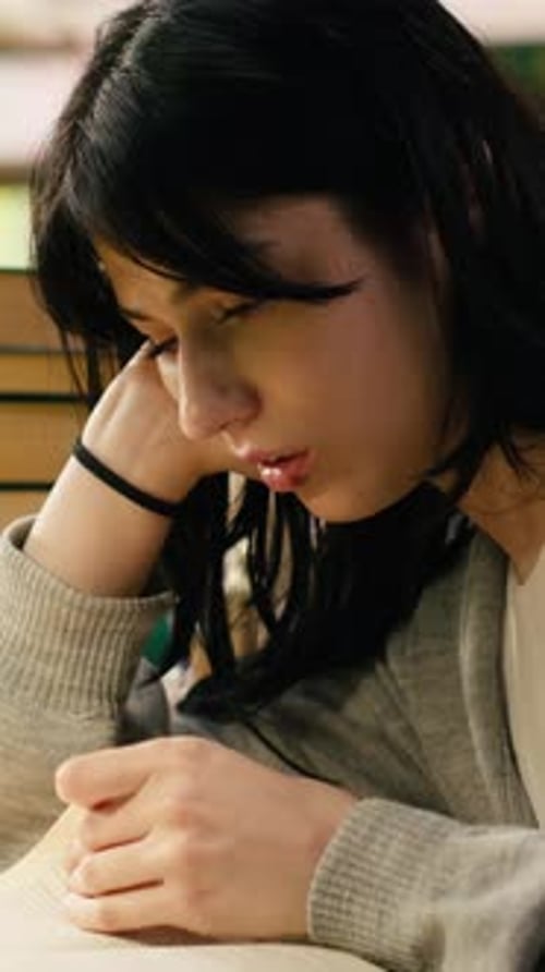 Vertical Video Tired Student Reading at Busy Library Table with Stacks of Books and Green Lamps