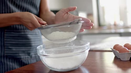 Midsection of biracial woman in apron sieving flour, baking in kitchen, slow motion