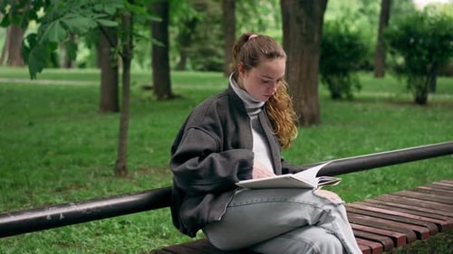 In the Park a Young Lonely Girl in a Black Jacket with Her Hair Pulled Together is Sitting on a