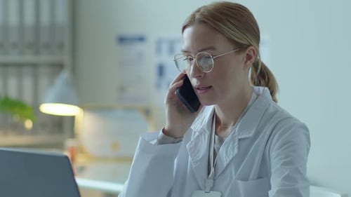 Female Doctor Using Laptop and Having Talk on Mobile Phone in Clinic