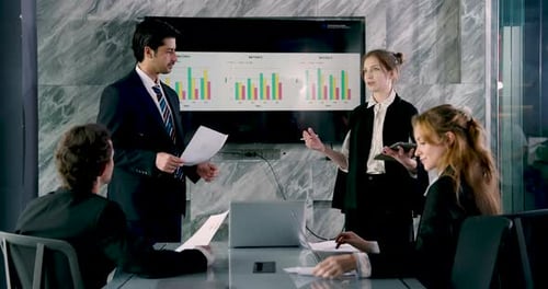 Male executive in formal suit presenting bar graph to colleagues in conference room, team of