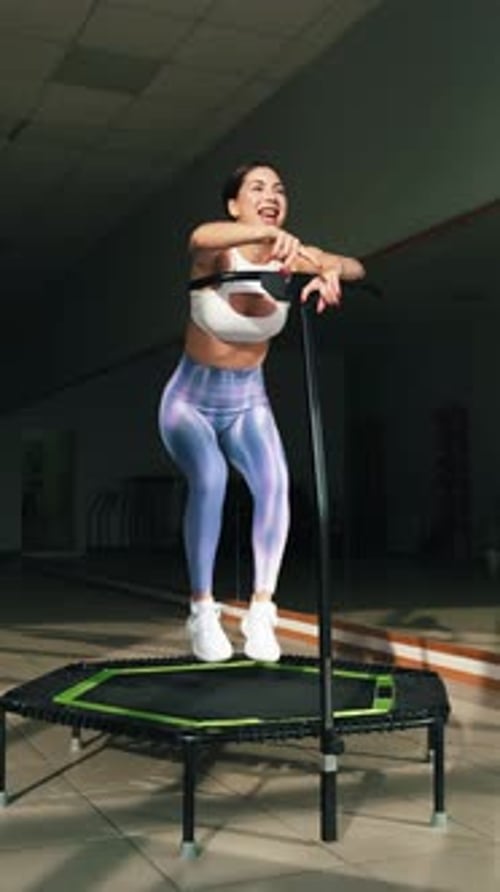 Joven y enérgica chica de fitness con top blanco y leggings azules iridiscentes haciendo ejercicios saltando sobre mo