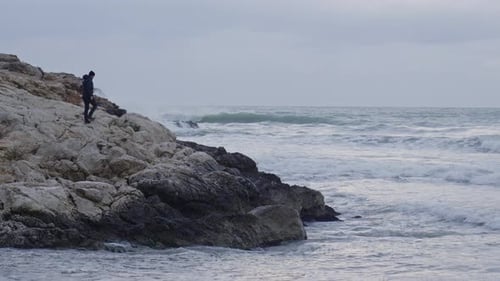 Man walks on rocks towards wild ocean front, kneels down and looks out towards stormy waves crushing