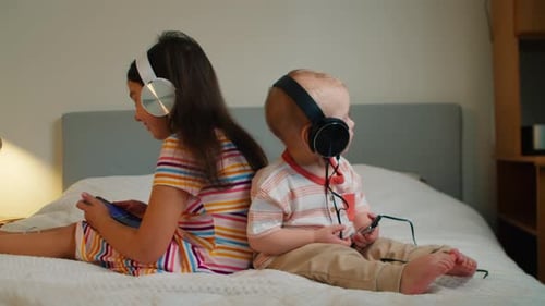 Siblings Using Smartphones with Headphones on Bed