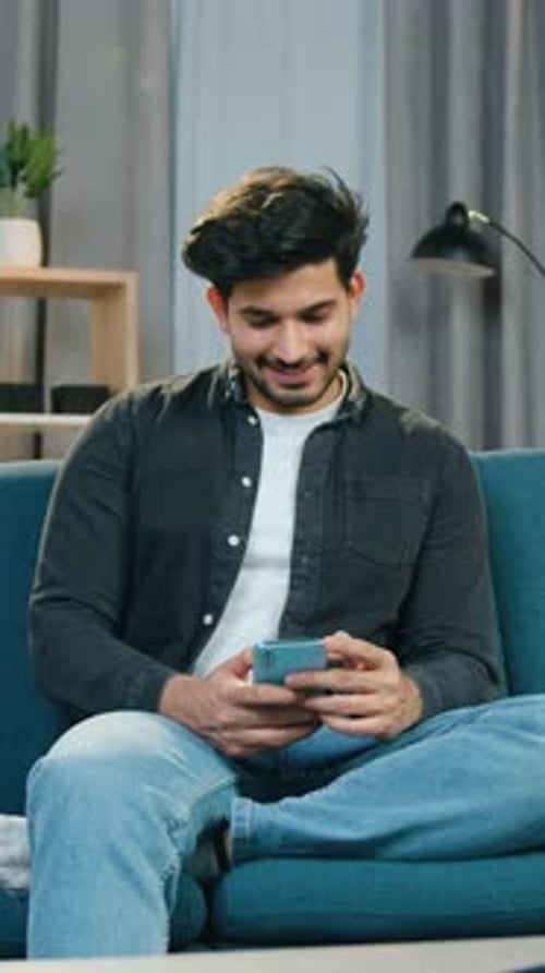 Smiling Young Man Using Smartphone on Couch Indoors