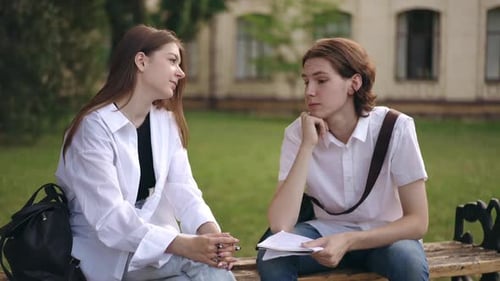 A Guy and a Girl are Sitting on a Bench on a Summer Day Outdoors Against the Backdrop of a Green