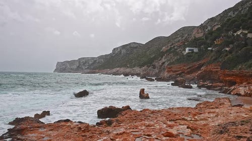 Coast in a bad weather day, gray sky, blue waves crashing into orange rocks of the coastline.