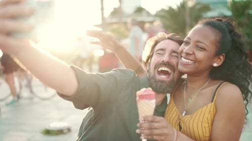 Closeup, smiling interracial couple eating ice cream in background of an urban city taking a selfie