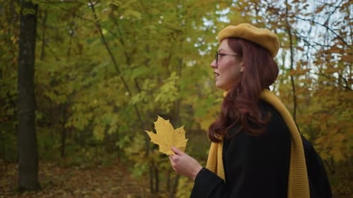 Smiling Woman Observing Autumn Leaf While Walking Through Forest in Fall