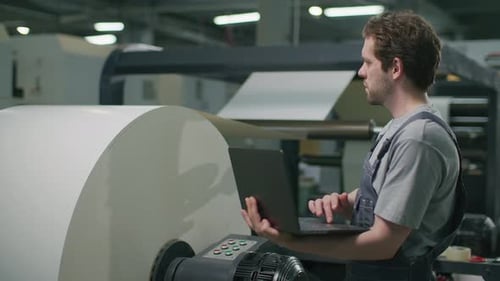 Industrial Operator Using Laptop to Monitor Web-Fed Press in Printing Factory