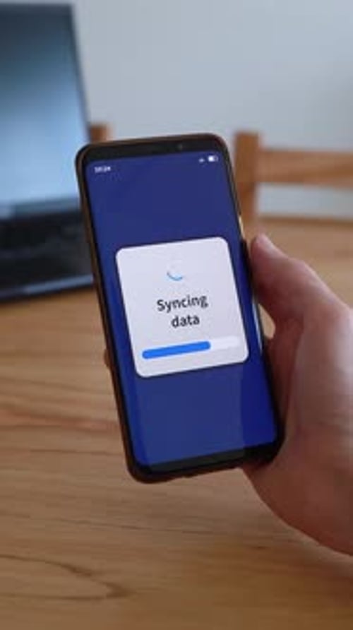 Vertical shot of smartphone syncing data with cloud storage. Laptop on the background.