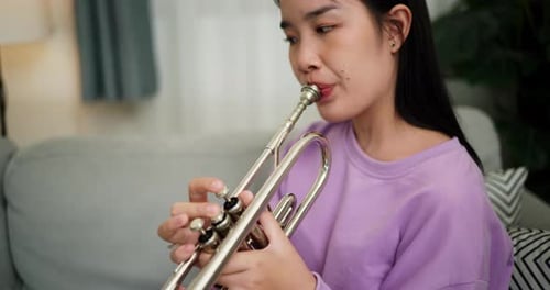 A young woman practices playing the trumpet while sitting on a sofa in a cozy living room.