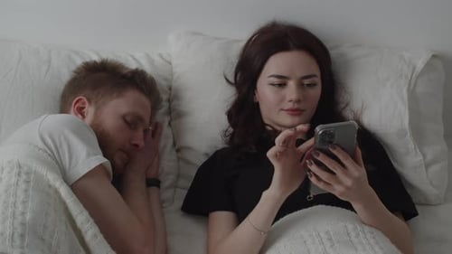 Couple in Bed, One Sleeping, One Using Phone