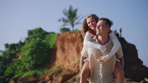 Happy Husband Carrying Wife Piggyback Running Barefoot on Tropical Sandy Beach Smiling Man and Woman