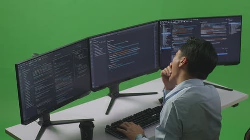 Side View Of Tired Asian Man Yawning While Write Code With Multiple Computer Screens In Green Screen