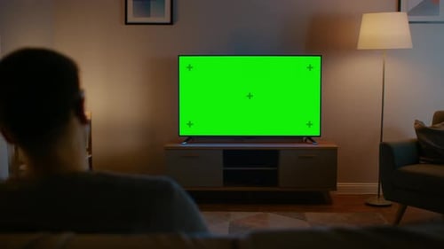 Young Man in Glasses is Sitting on a Sofa and Watching TV with Horizontal Green Screen Mock Up. It'