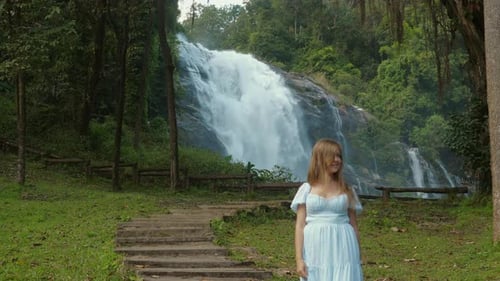 Woman in Blue Dress Standing Near Waterfall in Lush Green Forest