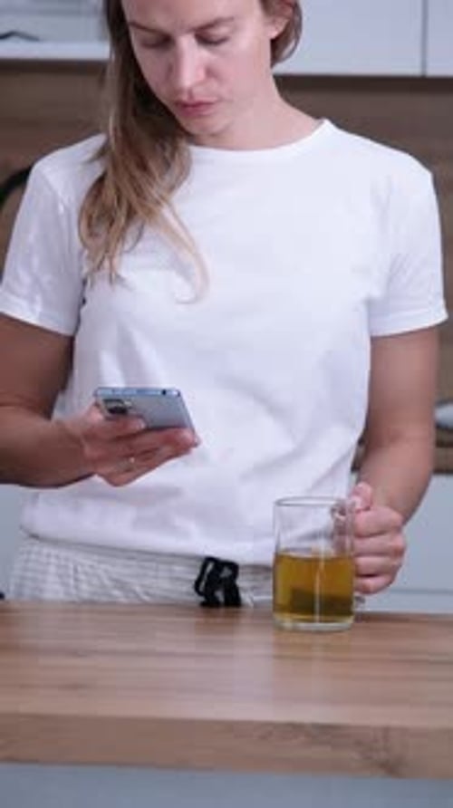 Woman Drinks Tea While Using Smartphone in Kitchen