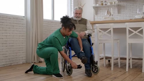 Woman Adjusting Wheelchair for Senior Man in Kitchen