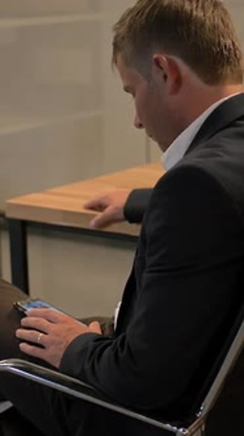 Vertical of Young Handsome Businessman Looking at His Smartphone Sitting on Chair in the Office