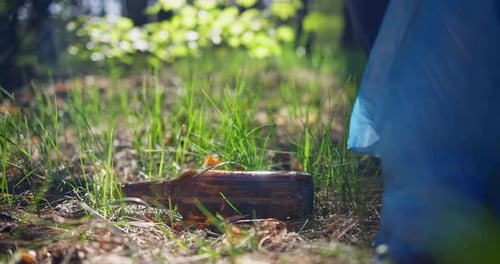 Un hombre con una bolsa de plástico recoge una botella de vidrio que está tirada en el césped mientras recoge basura en el