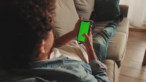 Young man lying on sofa uses mobile phone with green mock-up screen chroma key