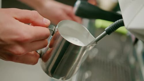 barista uses coffee machine and churns fresh milk to prepare latte or cappuccino in cafe for client