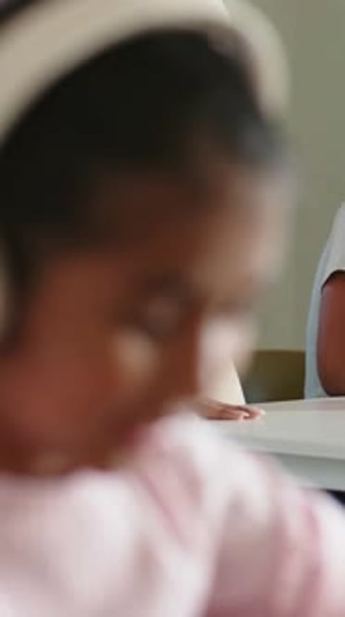 Vertical video: Camera refocusing girl in white headphones reading on device at table pink sweater